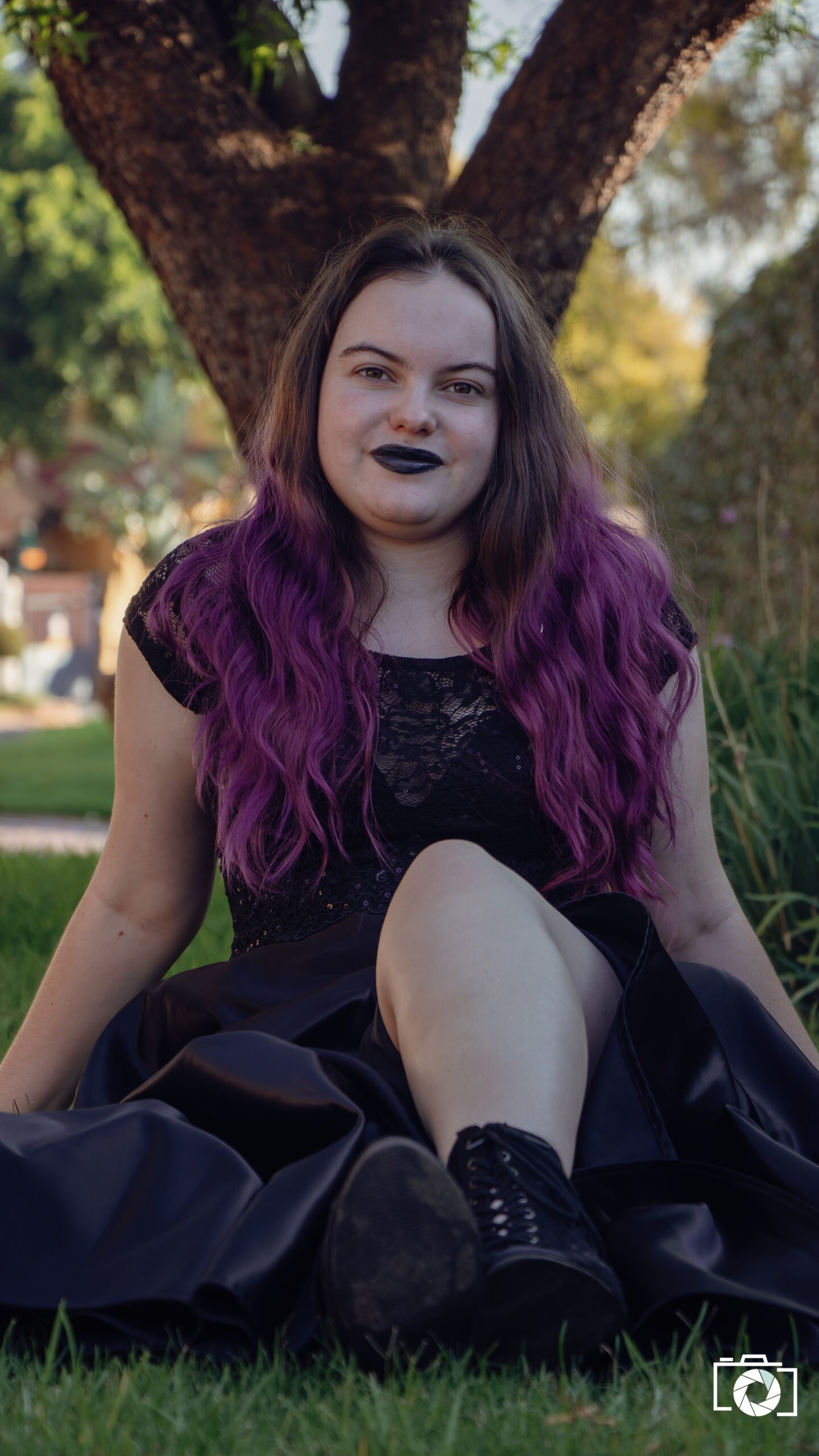 Young woman with purple hair sitting on grass, wearing a black dress, outdoors near a tree.