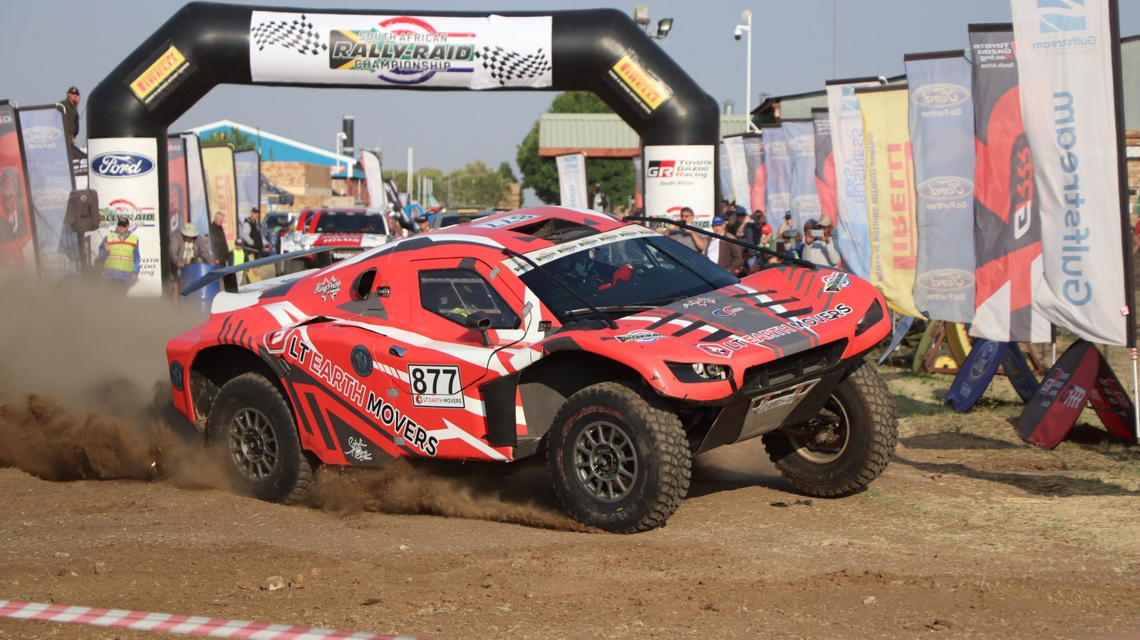 Red rally car racing at an event, kicking up dirt, with banners and spectators in the background.